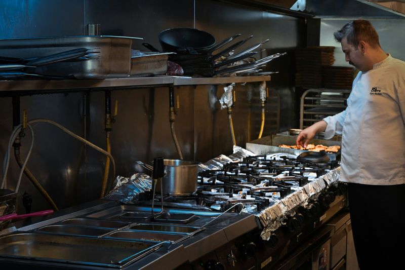 Chef in a commercial kitchen plating food at a gas range, surrounded by pots, pans and stainless steel prep equipment