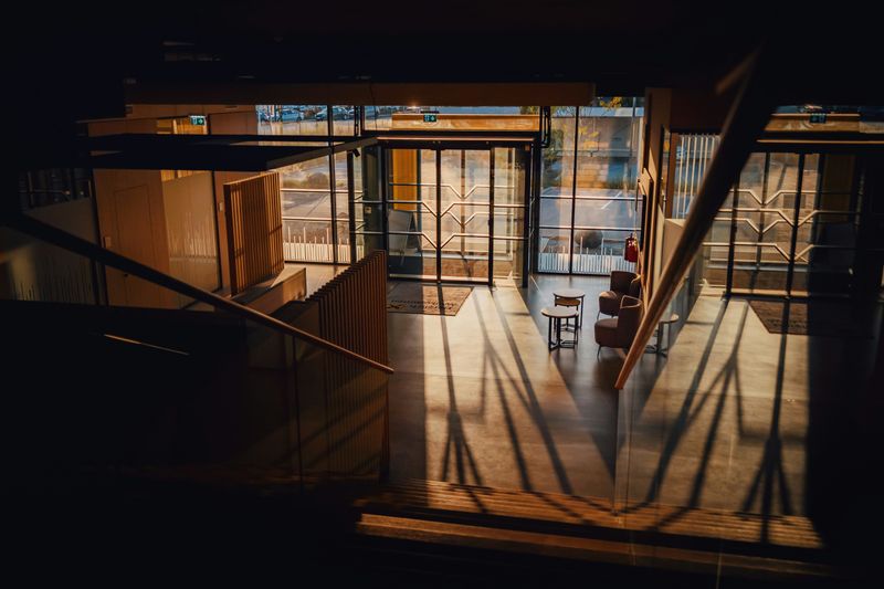 Sunlit office building lobby seen from an upper mezzanine, with glass sliding entrance doors, timber slat panelling, concrete flooring and lounge seating below
