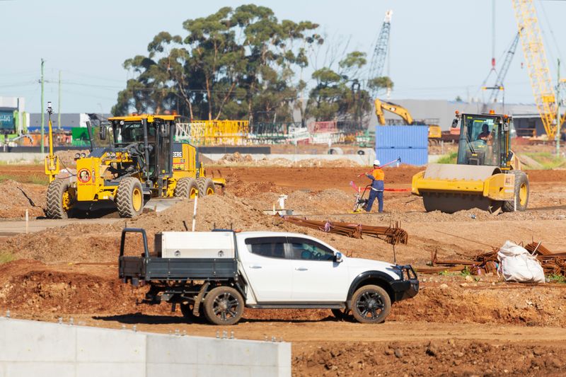 Active construction site with a yellow road grader, roller and excavator, a white tradesman's ute, and a worker in high-visibility clothing operating a concrete screed among reinforcement steel bars